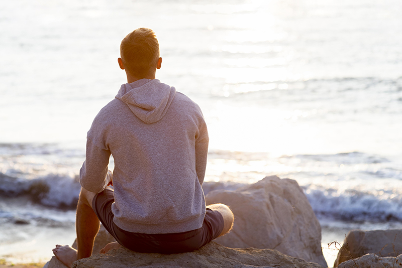Man zit op een steen terwijl hij naar de zee kijkt bij zonsondergang en doet aan mindfulness