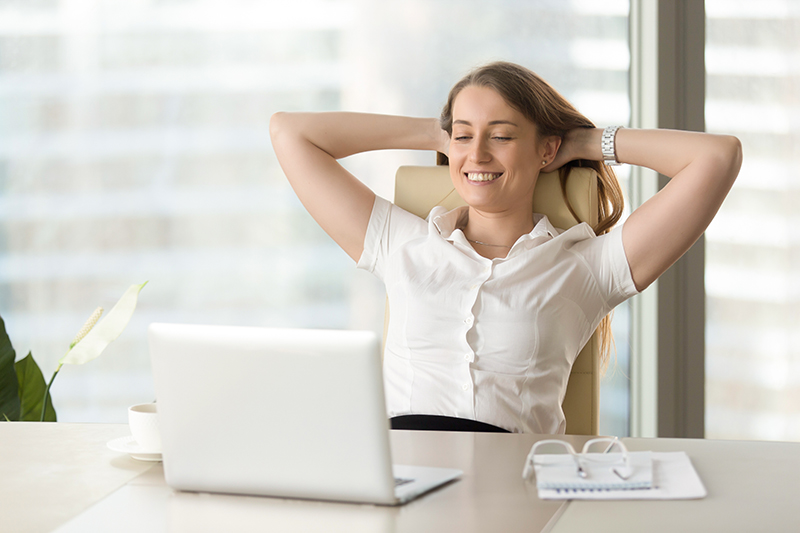 Vrouw voorkomt hoge rugpijn en zit op een stoel aan haar bureau terwijl ze glimlacht en tevreden is met haar werk.