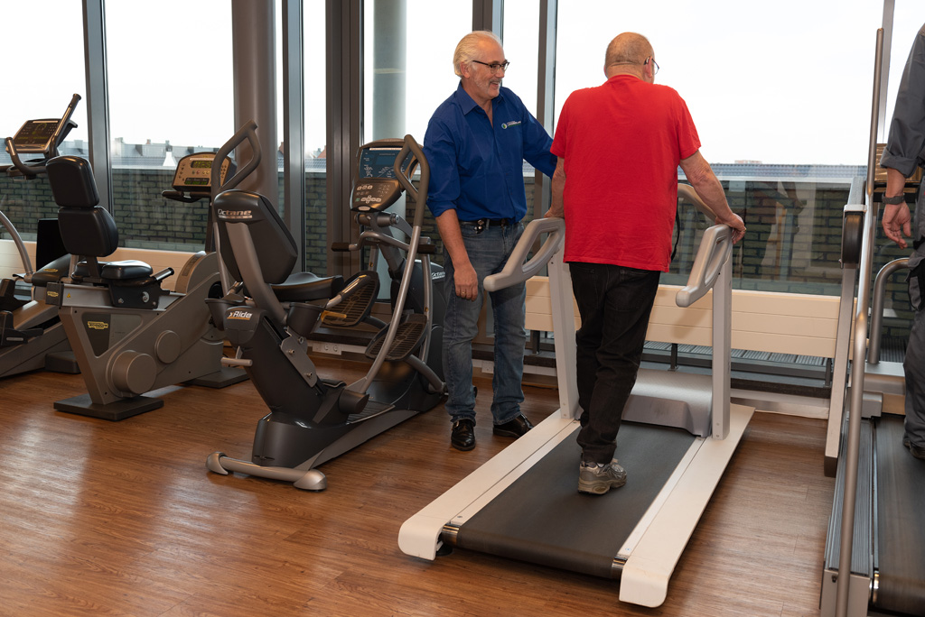 Man in een rood shirt met artritis loopt op de loopband naast een fysiotherapeut met blauw shirt aan in de oefenzaal.
