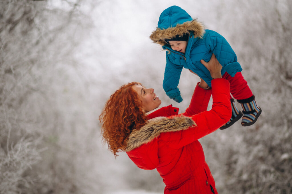 vrouw in een rode jas heeft geen last van rugpijn in de winter en tilt haar zoontje in een blauw jas op in de lucht