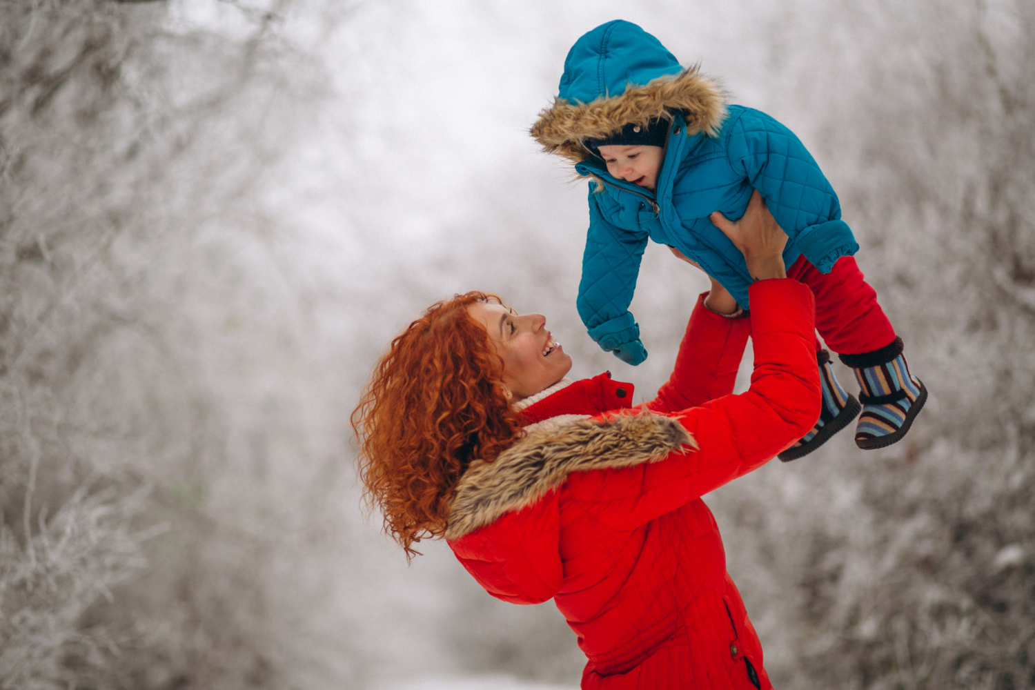 Rugpijn in de winter – Lansingerland vrouw in een rode jas heeft geen last van rugpijn in de winter en tilt haar zoontje in een blauw jas op in de lucht