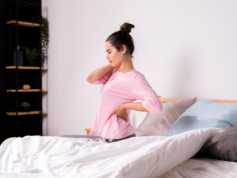 Een vrouw in een roze pyjama zit op een bed en heeft last van aspecifieke rugklachten.