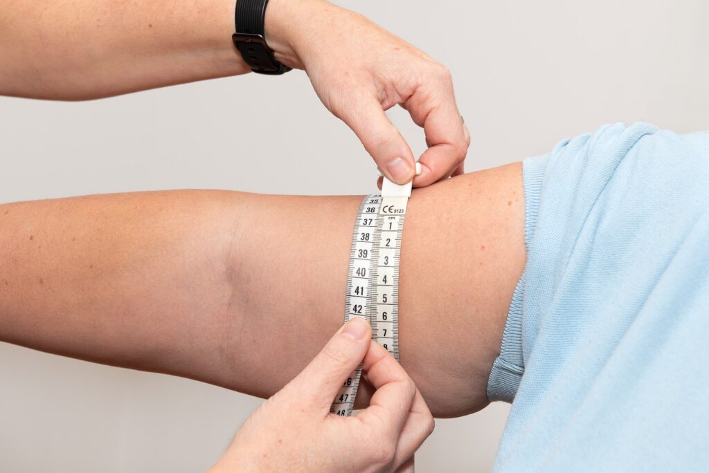 Een patiënt met een lichtblauw t-shirt steekt zijn arm uit en de therapeut met een zwart horloge meet zijn arm met een meetlint.