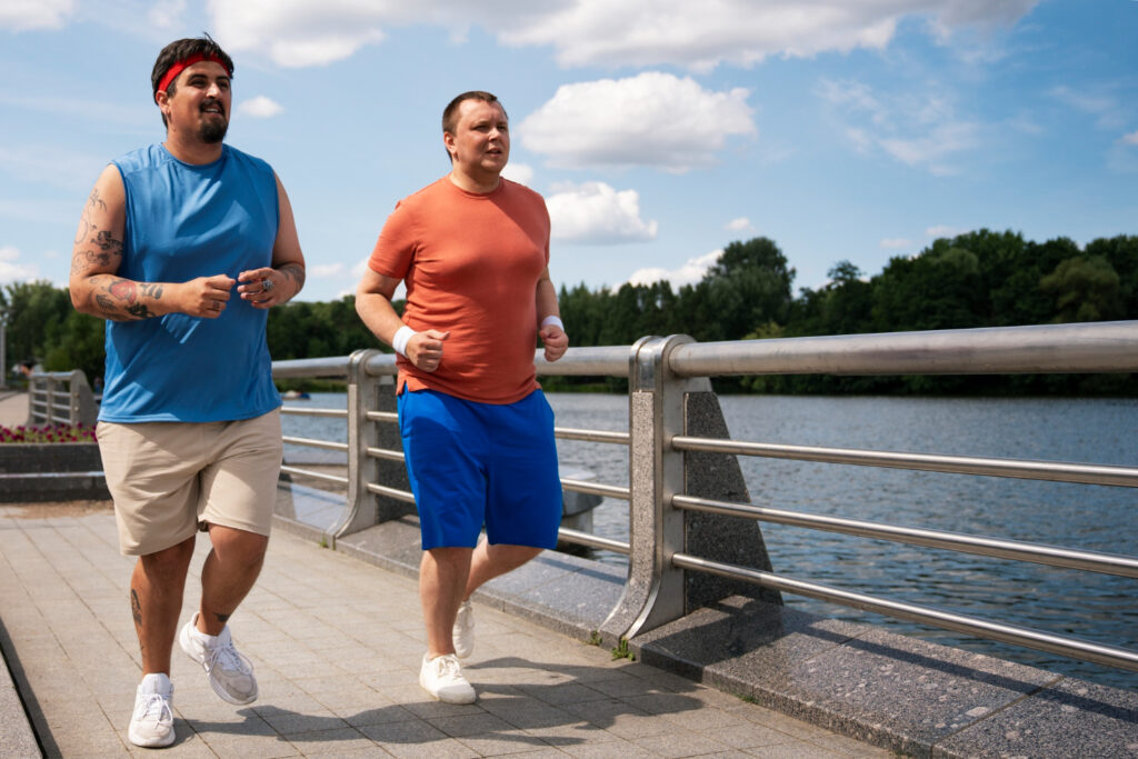 Twee personen met overgewicht in een blauw shirt en roze shirt rennen over de brug boven het water en werken werken aan hun obesitas door middel van een programma met GLI