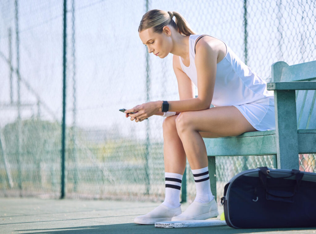 Sportster in tenniskleding is bezig met haar Polsklachten door smartphonegebruik en zit buiten op de bank.